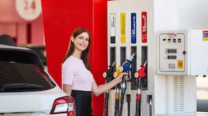Person refuelling a white car at a Shell service station, holding a fuel nozzle beside pumps displaying Shell fuel grades including Unleaded 91, Unleaded 95 and Shell V-Power.