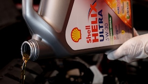 Close-up of Shell Helix Ultra 5W-30 motor oil being poured from a container into a vehicle engine by a person wearing protective gloves.