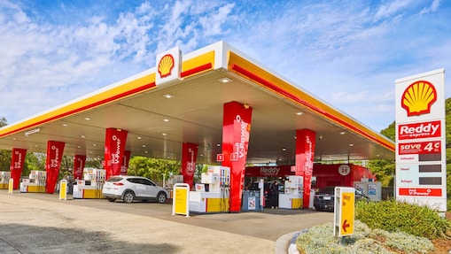 Wide exterior view of a Shell Reddy Express service station with multiple fuel pumps, vehicles, Shell branding and price signage under a large canopy.