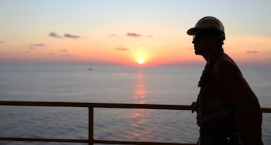 A worker wearing PPE holds a handrail and looks out over the ocean at sunrise.
