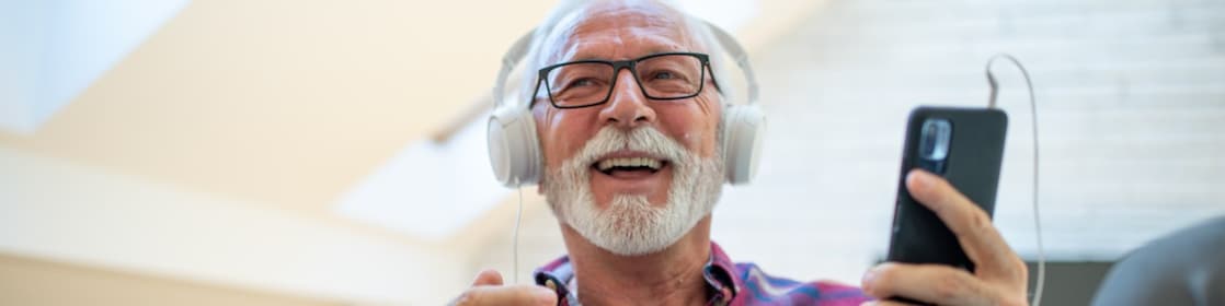 A smiling older man with a white beard and glasses holds a smartphone connected to his headphones.