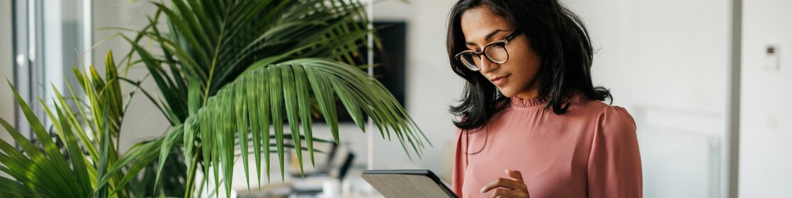 A woman stands in an empty office looking at the tablet she is holding.