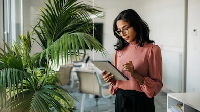 A woman stands in an empty office looking at the tablet she is holding.