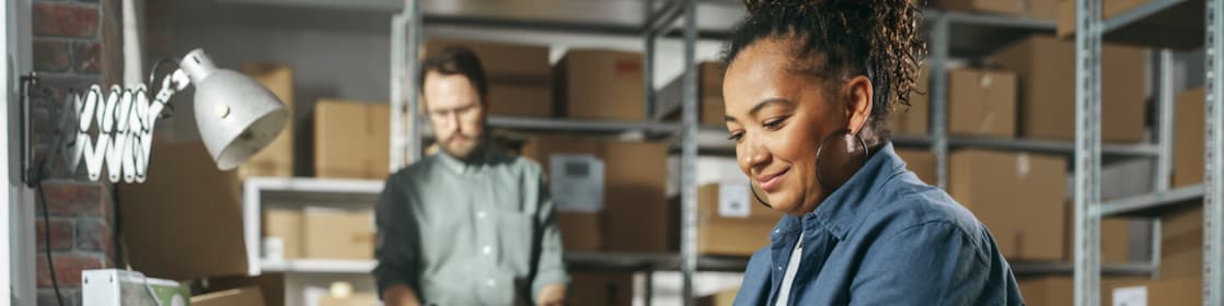 Diverse Male and Female Warehouse Inventory Managers Talking, Using Laptop Computer and Checking Retail Stock. Rows of Shelves Full of Cardboard Box Packages in the Background.