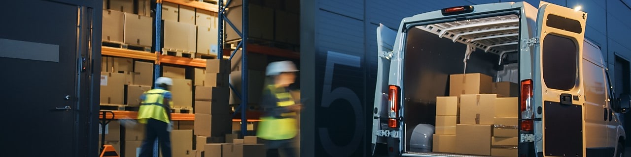 Two workers wearing hi-vis move boxes from a van stopped outside a warehouse.