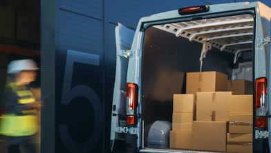 Two workers wearing hi-vis move boxes from a van stopped outside a warehouse.