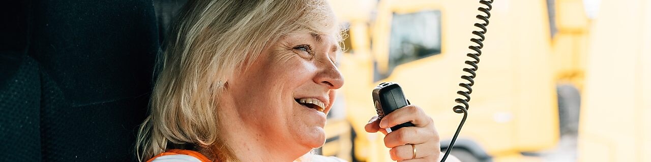 A woman in a high-visibility vest smiles as she holds a CB radio in a large vehicle