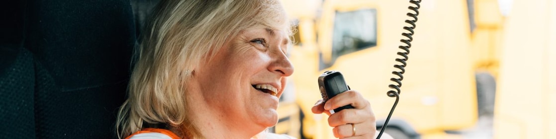 A woman in a high-visibility vest smiles as she holds a CB radio in a large vehicle