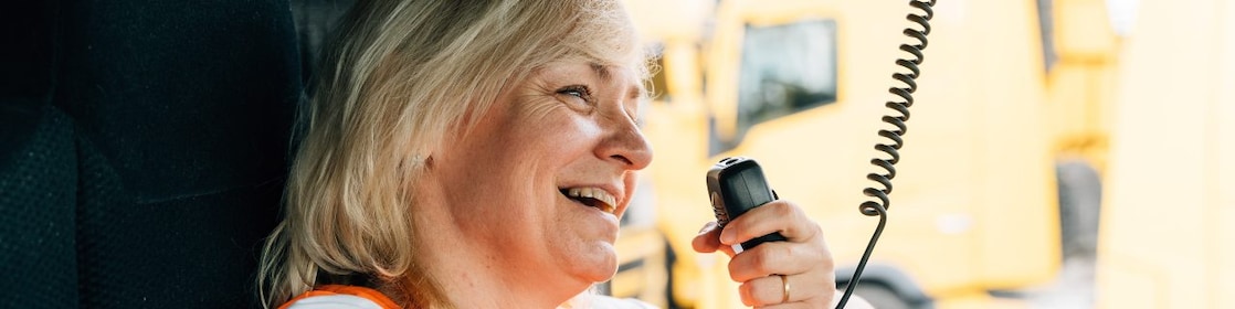 A woman in a high-visibility vest smiles as she holds a CB radio in a large vehicle