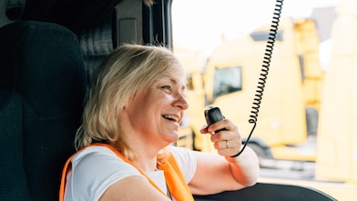 A woman in a high-visibility vest smiles as she holds a CB radio in a large vehicle
