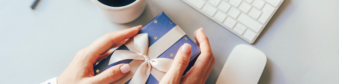Blue gift box decorated with a golden ribbon bow in female hands against the background of the office workplace, gift delivery, coffee break during office hours, corporate greetings and parties.