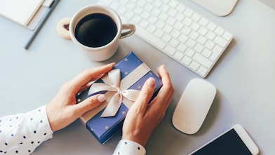 Blue gift box decorated with a golden ribbon bow in female hands against the background of the office workplace, gift delivery, coffee break during office hours, corporate greetings and parties.