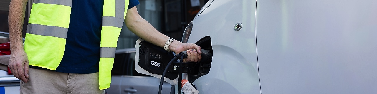 A man in a safety vest is positioned next to a white car, engaged in the process of charging an electric vehicle.