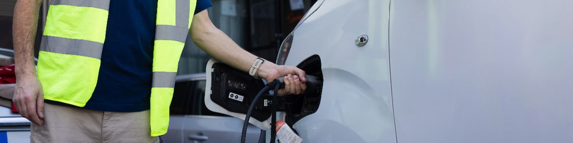 A man in a safety vest is positioned next to a white car, engaged in the process of charging an electric vehicle.
