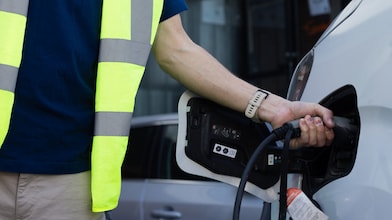 A man in a safety vest is positioned next to a white car, engaged in the process of charging an electric vehicle.