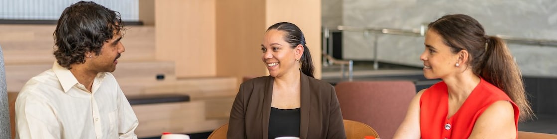 Three coworkers sit around a round black table in a contemporary office lounge, talking and holding coffee cups.