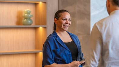 Person in a blue jacket holds a phone, speaking with a colleague in a modern office with lit wooden shelving.