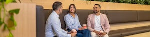 Three coworkers chat on bench seating, holding coffee cups in a modern office lounge with indoor plants.