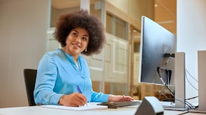 Person writing in a notebook at a desk, working at a computer in a modern glass‑walled office.