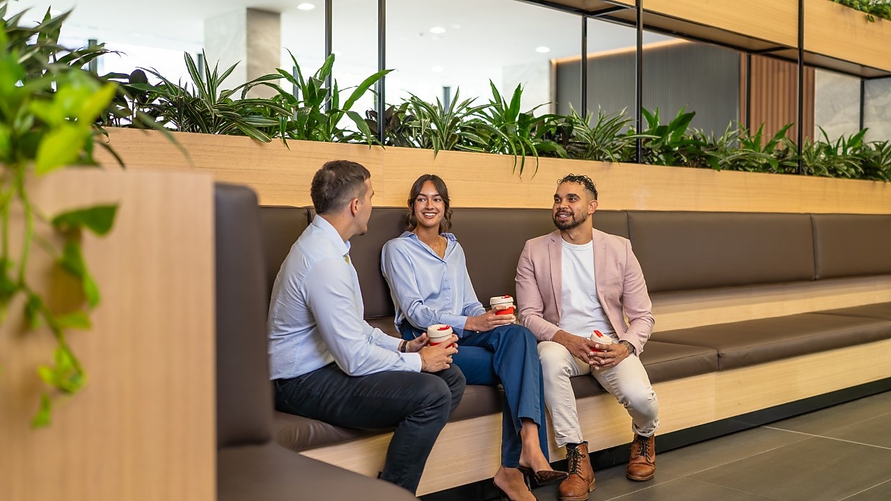 Three coworkers chat on bench seating, holding coffee cups in a modern office lounge with indoor plants.