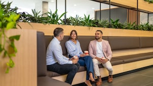 Three coworkers chat on bench seating, holding coffee cups in a modern office lounge with indoor plants.