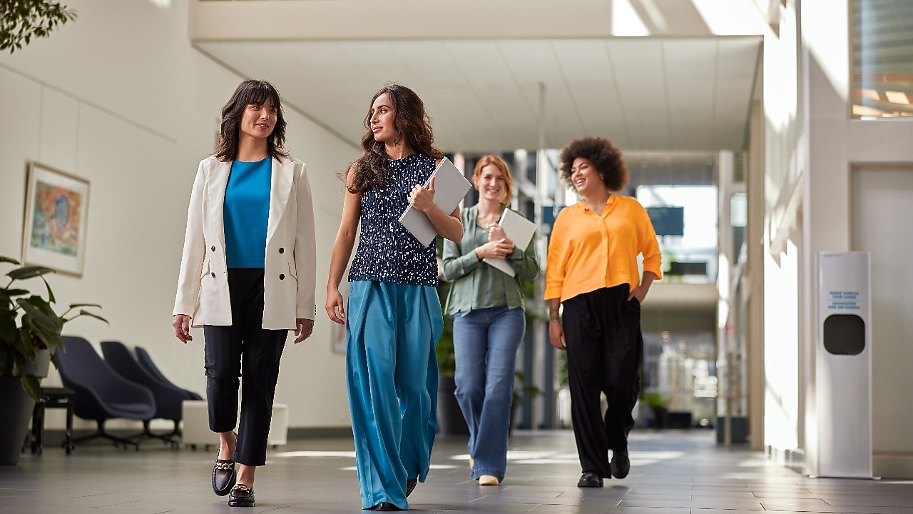 Four people walking through a bright office hallway, carrying notebooks and folders, with seating and artwork visible in the background.