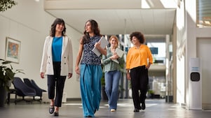 Four people walking through a bright office hallway, carrying notebooks and folders, with seating and artwork visible in the background.