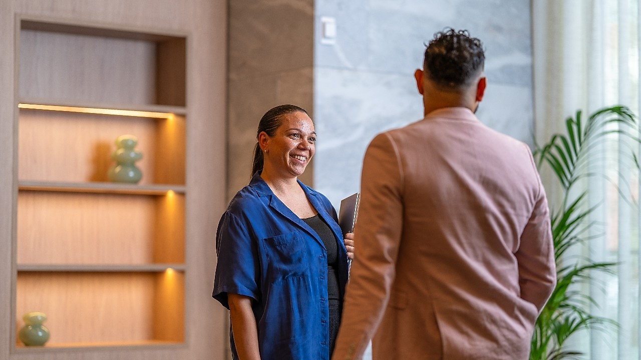 Two people standing indoors near a wooden shelf, engaged in conversation; one holds a folder in hand.