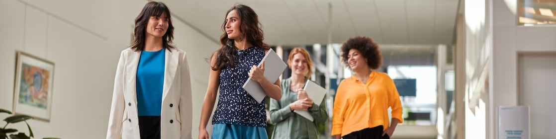Four people walking through a bright office hallway, carrying notebooks and folders, with seating and artwork visible in the background.