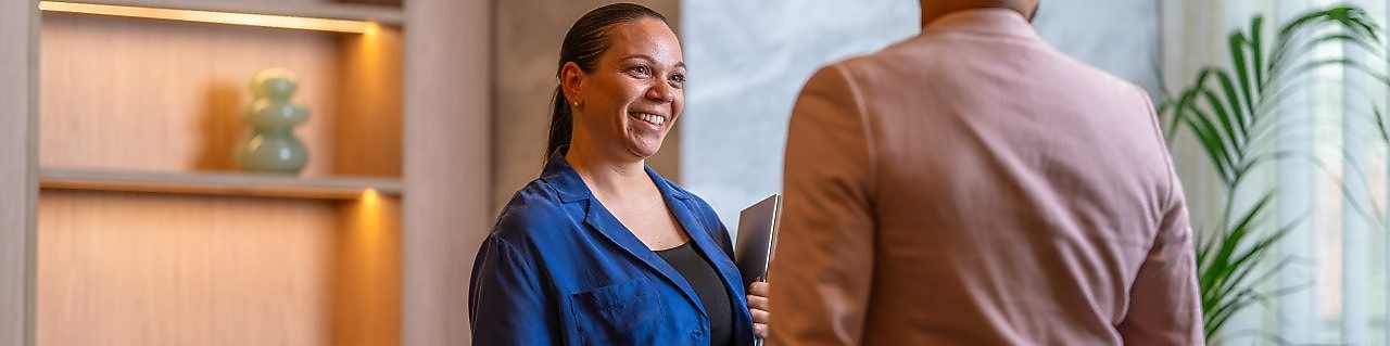 Two people standing indoors near a wooden shelf, engaged in conversation; one holds a folder in hand.
