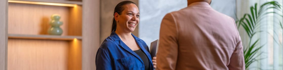 Two people standing indoors near a wooden shelf, engaged in conversation; one holds a folder in hand.