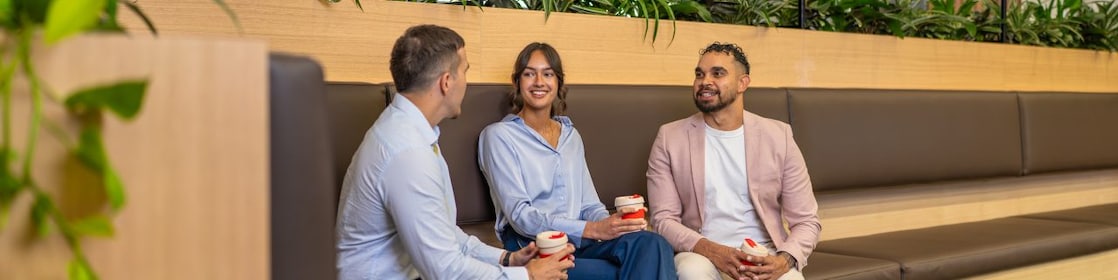 Three coworkers chat on bench seating, holding coffee cups in a modern office lounge with indoor plants.