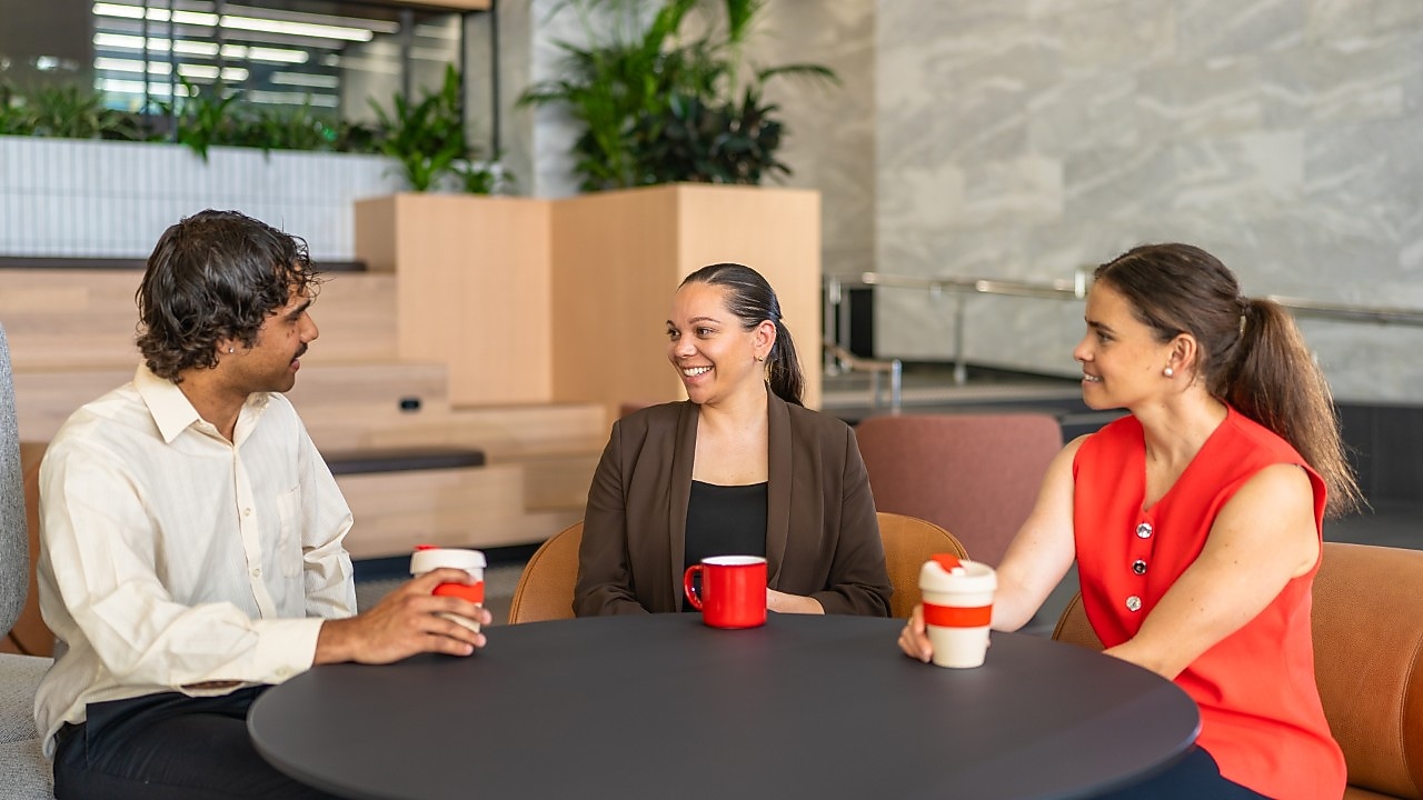 Three coworkers sit around a round black table in a contemporary office lounge, talking and holding coffee cups.