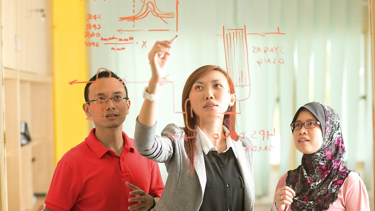 Colleagues gathered standing and discussing metrics on a glass board
