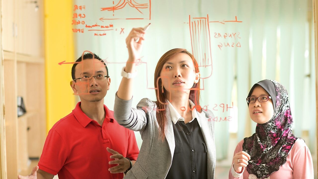 Colleagues gathered standing and discussing metrics on a glass board