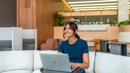 Person uses an HP laptop on a sofa in a modern office lounge with plants and wood‑slat walls.