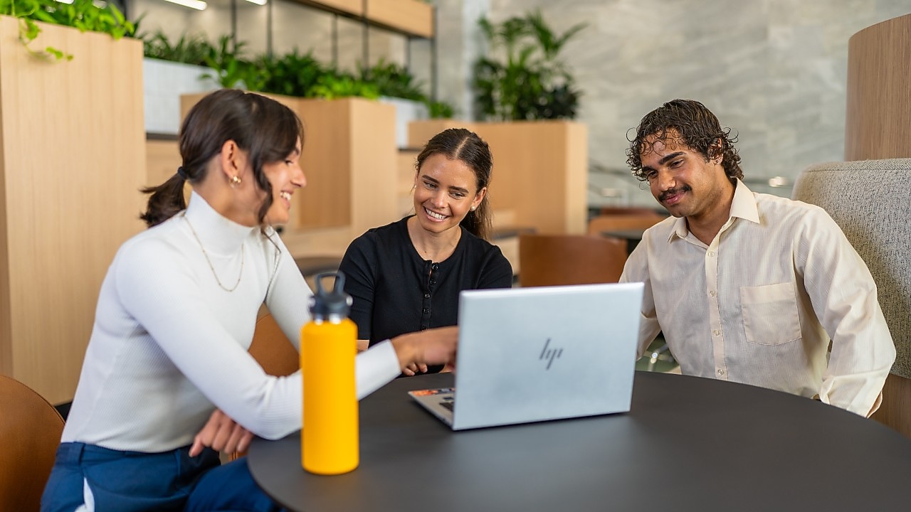 Three colleagues work together around a laptop at a round table in a modern office lounge with plants.