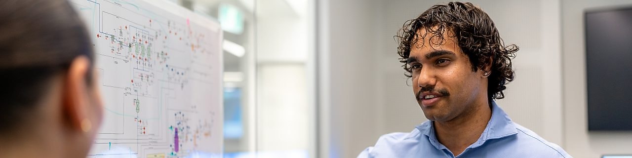 Engineer in a blue shirt points to a technical diagram on a whiteboard while explaining to a colleague.