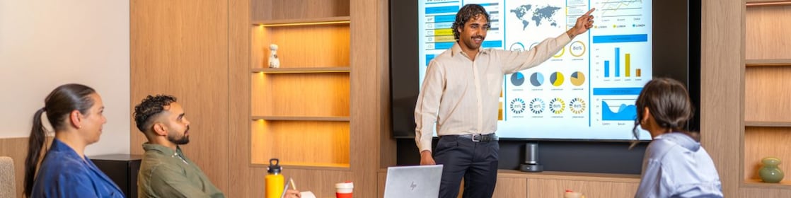 Presenter points to charts on a large screen; three colleagues watch with laptops and notebooks in a meeting room.