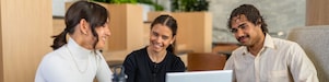 Three colleagues work together around laptop at a round table in a modern office lounge with plants.