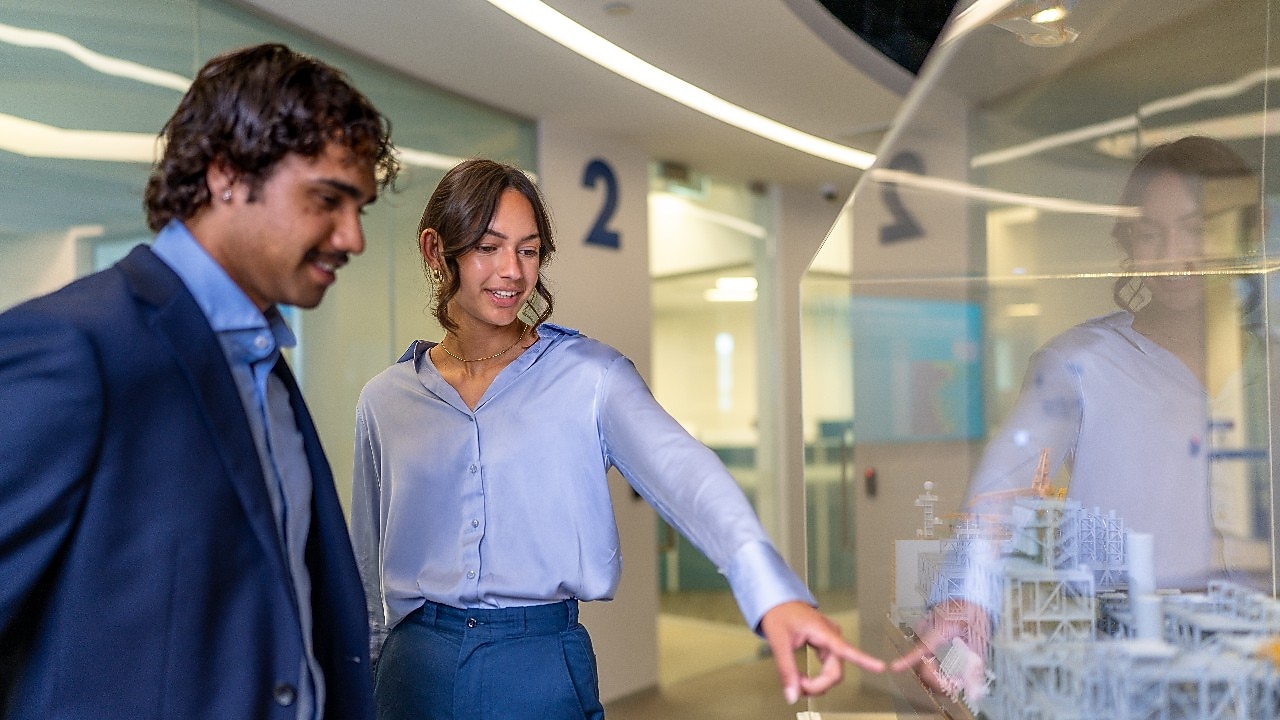 Two individuals in a modern office environment standing near a glass display case. One person is pointing at a detailed scale model of industrial structures inside the case, while the other observes. The background shows numbered wall sections and bright lighting.