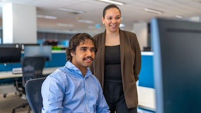 Two individuals in a modern office setting, with one seated at a desk working on a computer and the other standing nearby. The workspace features multiple monitors, chairs, and blue partition panels in the background.