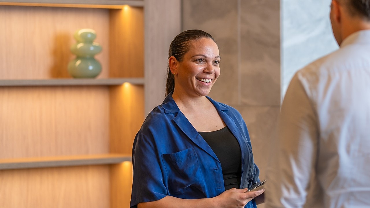 person in a blue jacket holds a phone, speaking with a colleague in a modern office with lit wooden shelving