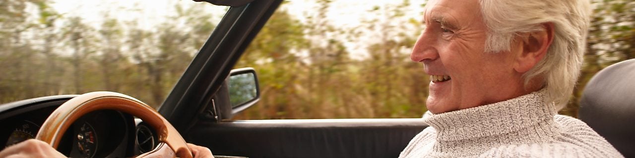 A man drives an open-top car along a tree-lined road