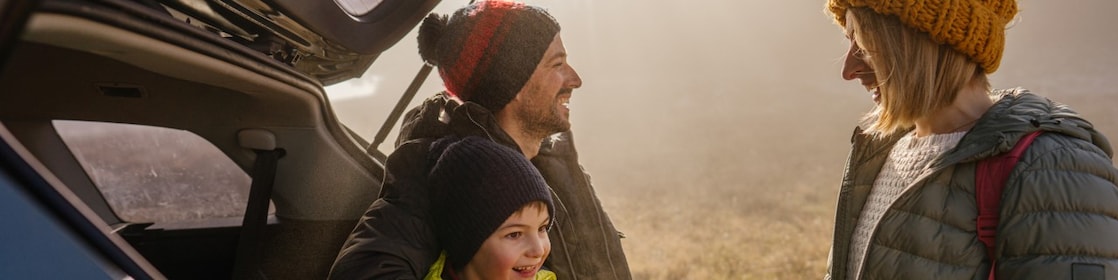 A young family of three stand by the boot of their 4WD wearing beanies and winter clothing, parked by a foggy field.