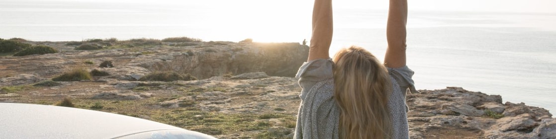 A woman stands by her car, stretching her arms in the air