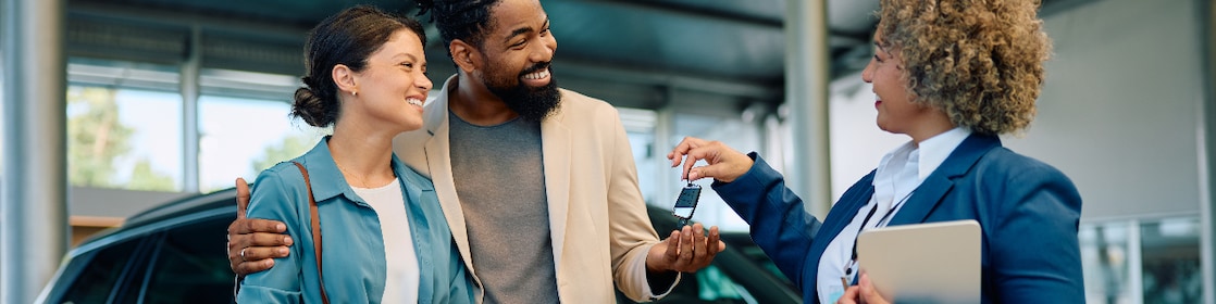 Happy multiracial couple receiving keys of their new car from saleswoman in showroom.