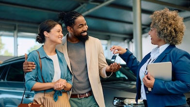 Happy multiracial couple receiving keys of their new car from saleswoman in showroom.