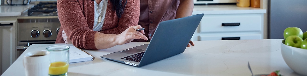 Caucasian woman and African American man sit at kitchen counter with breakfast working with pen, paper and laptop.