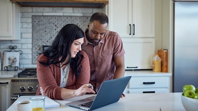 Caucasian woman and African American man sit at kitchen counter with breakfast working with pen, paper and laptop.